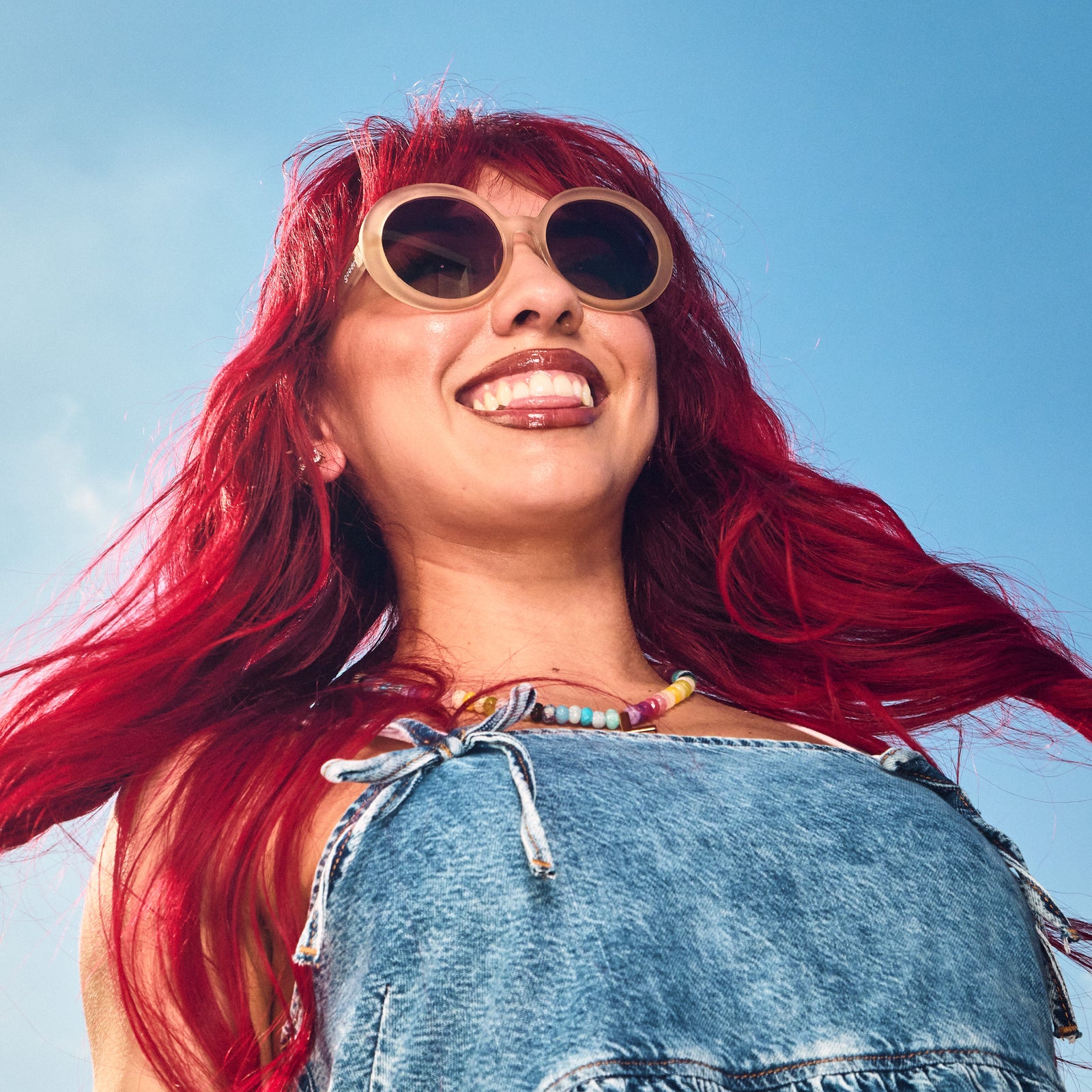 Low-angle view of a red-headed woman with oversized, round beige sunglasses with purple gradient lenses.