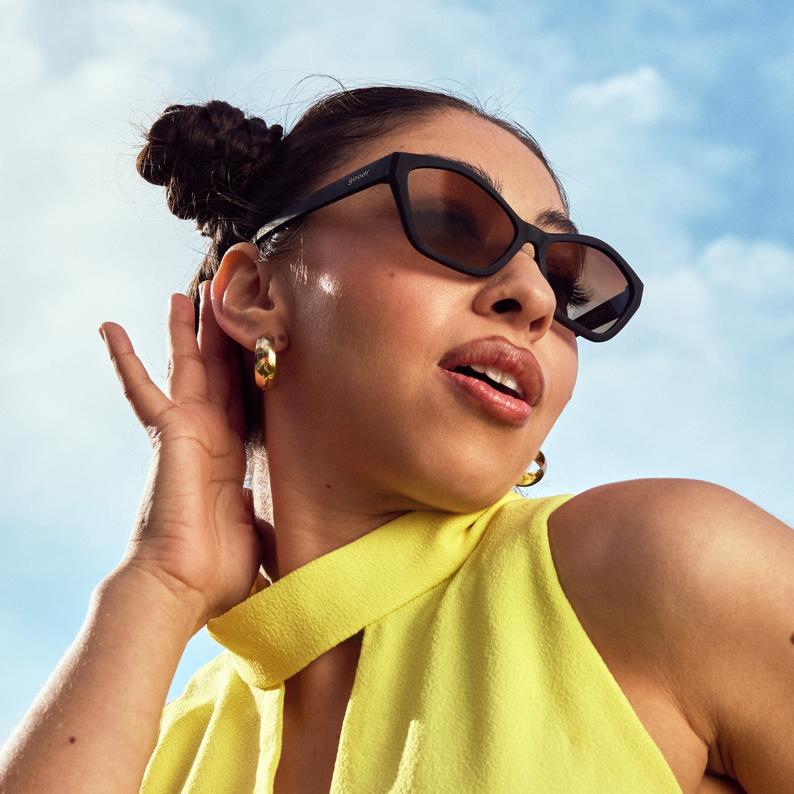 Low-angle view of a woman posing, wearing black geometric cat-eye sunglasses with amber gradient lenses.