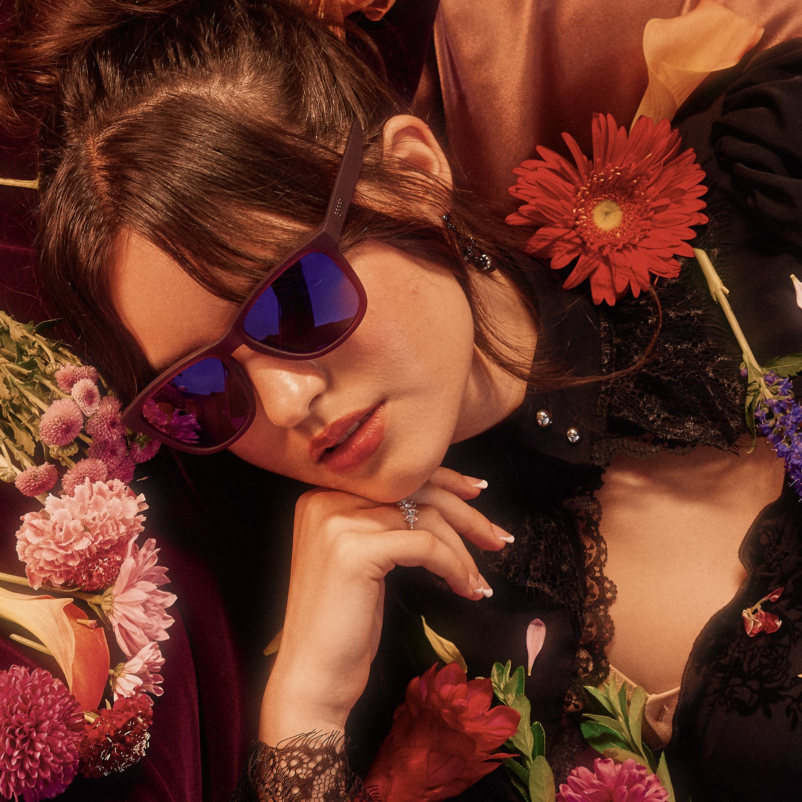 A woman surrounded by flowers, poses while wearing translucent purple sunglasses with reflective purple lenses.