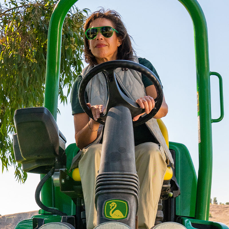 A woman wearing green aviator sunglasses with non-reflective lenses driving a green tractor.