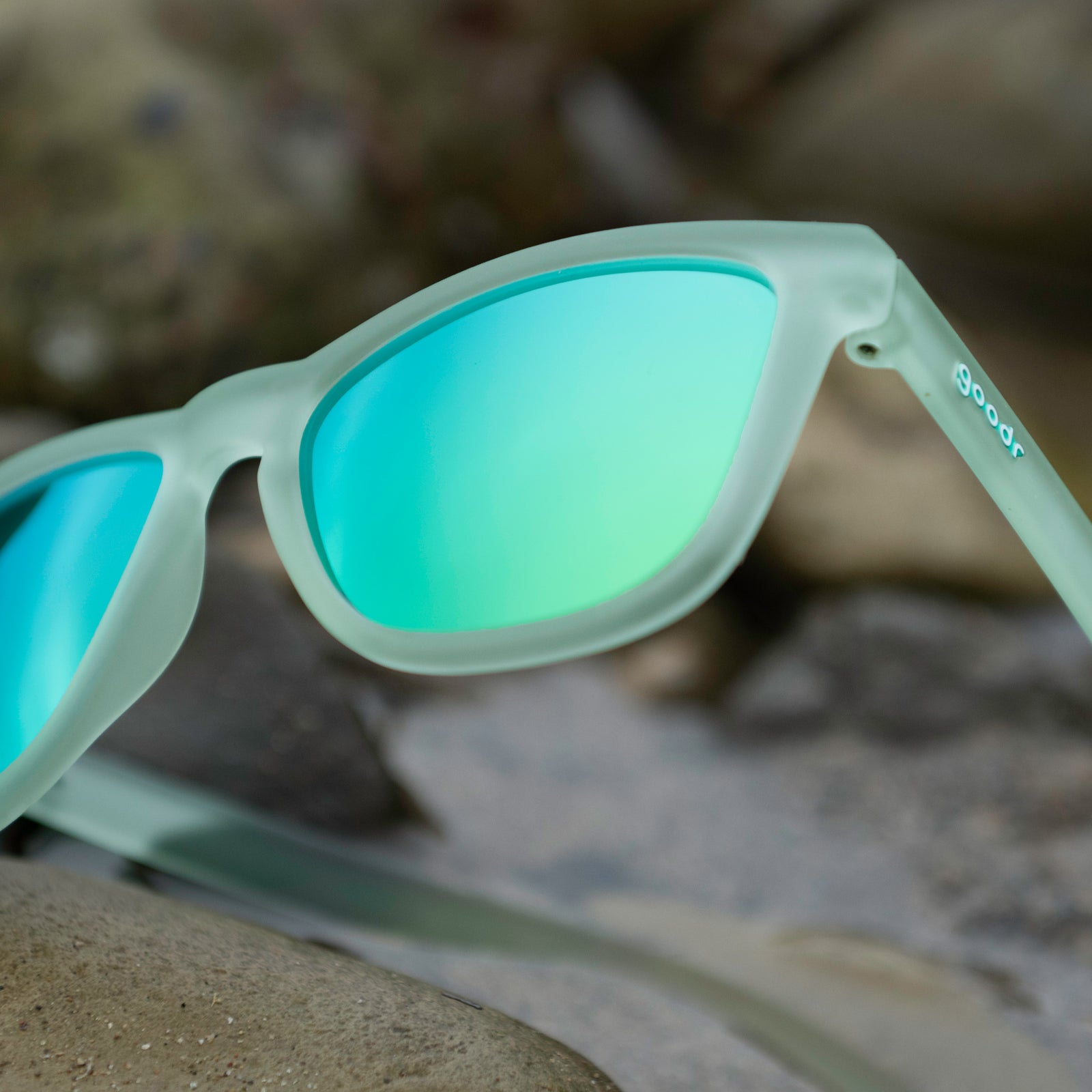 Close-up of translucent green OG sunglasses with green-blue reflective lenses with sea rocks in the background.