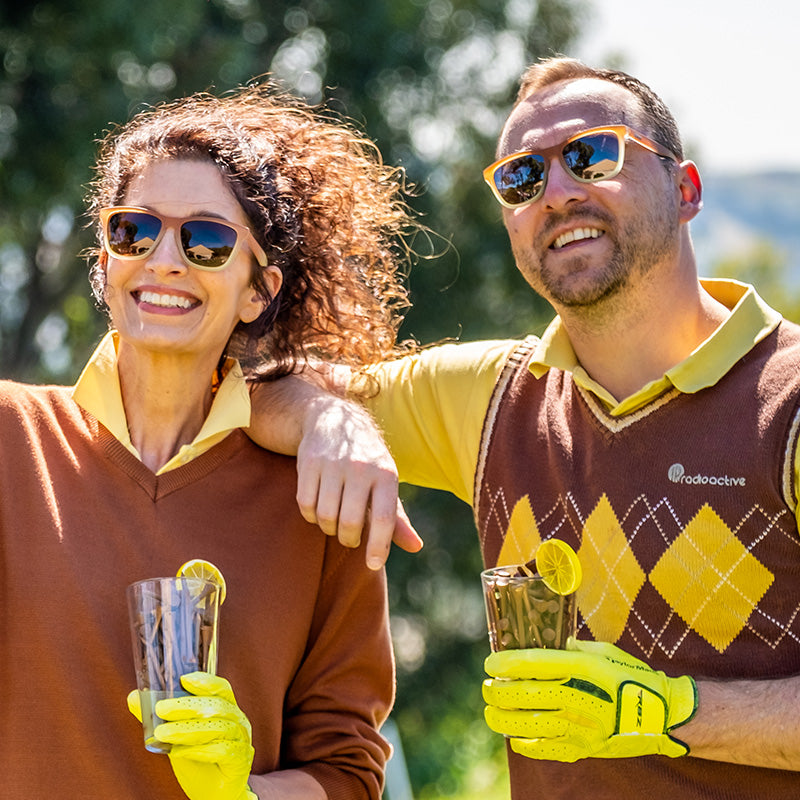 A man and woman wearing brown-to-white gradient sunglasses with brown lenses smile on a golf course holding iced tea.
