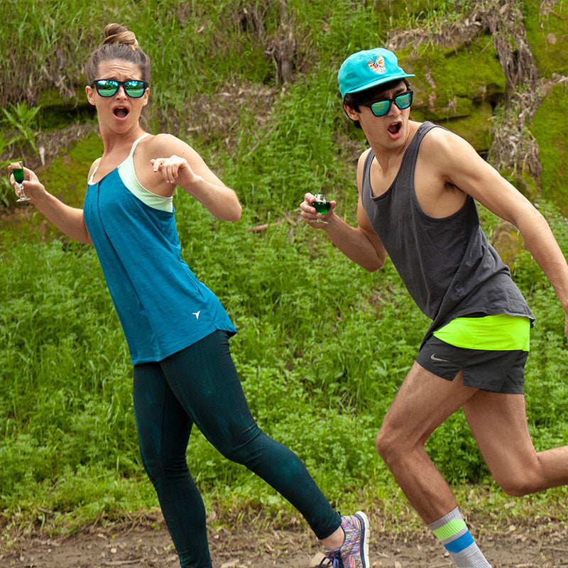 Two runners wearing black sunglasses with green reflective lenses look over their shoulders, afraid as they jog in a meadow.