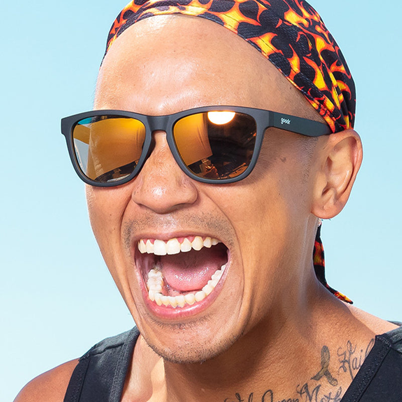 A man wearing a flame-patterned durag and black sunglasses with amber reflective lenses gleefully shouts.
