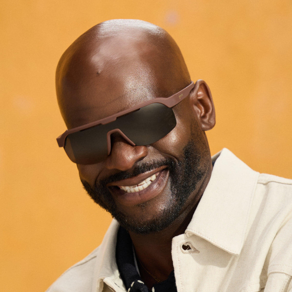 A man smiles confidently, wearing a pair of brown wraparound Bolt G sunglasses against a golden, ochre-yellow background.