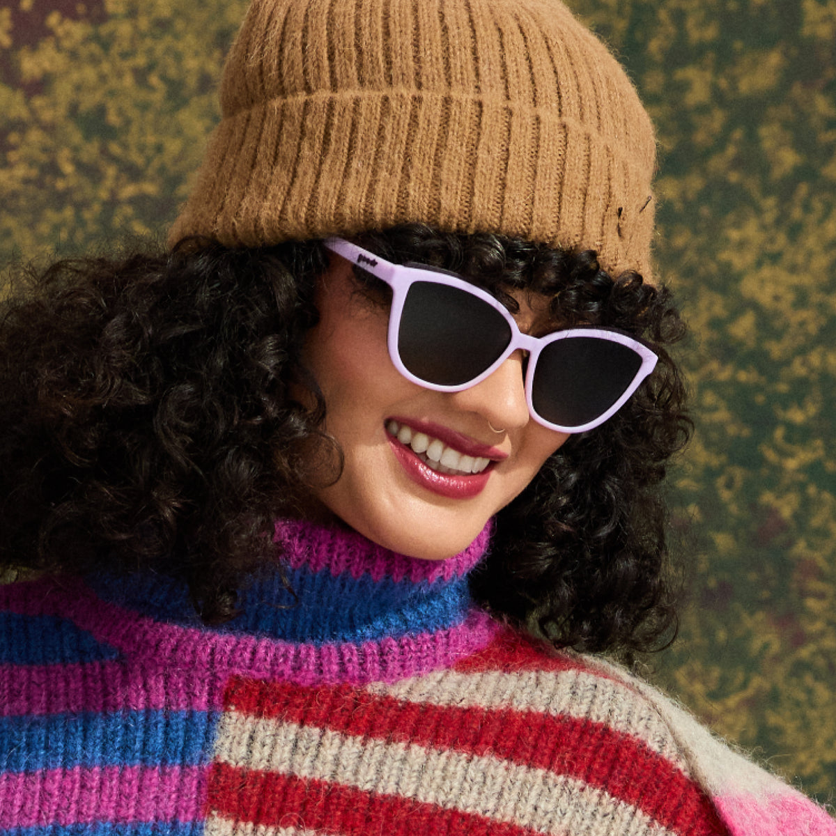 A dark-haired woman in a camel-colored beanie and colorful fuzzy striped sweater smiles in a pair of lavender Pounce Gs.