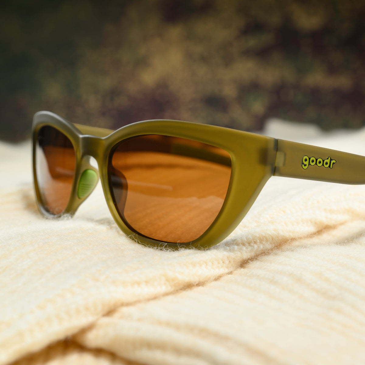 A pair of translucent olive-green cat-eye sunglasses with brown non-reflective lenses sits on a cream-colored sweater.