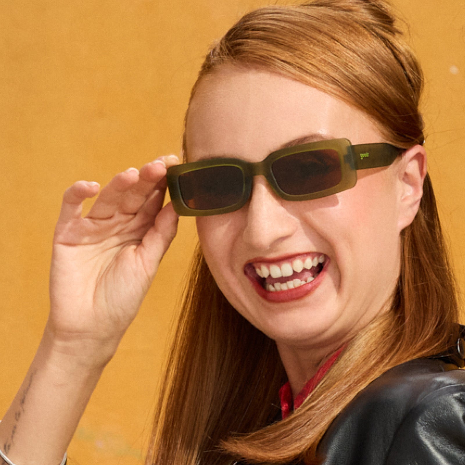 A woman with long red hair smiles in a pair of translucent, rectangular olive-green sunglasses with brown lenses.