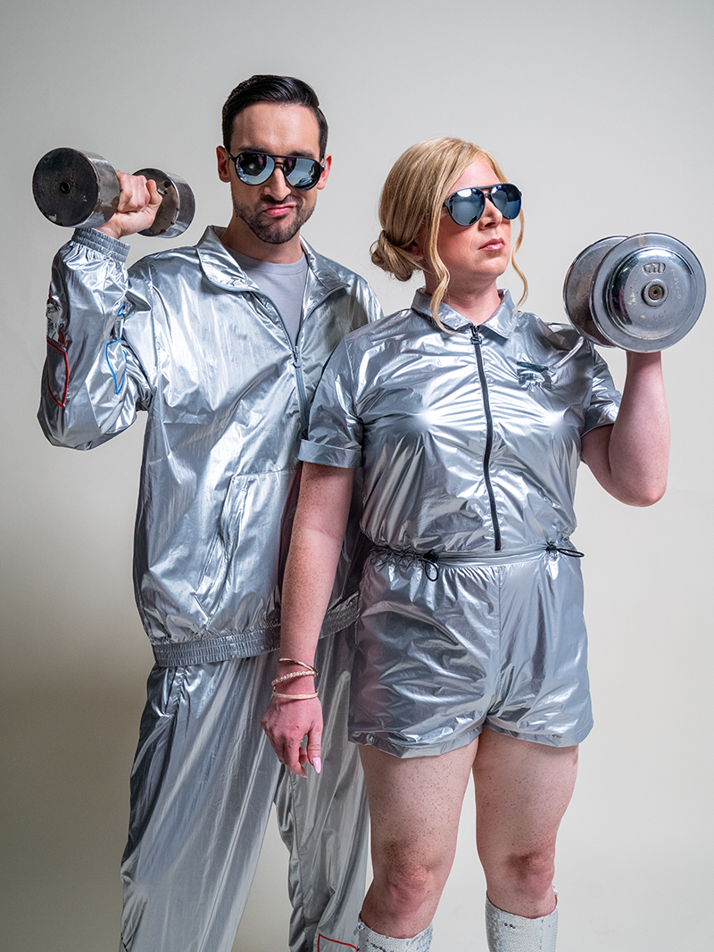 A man and a woman wearing aviator sunglasses with black frames and reflective lenses, in metallic silver jumpsuits, holding metal dumbbells.