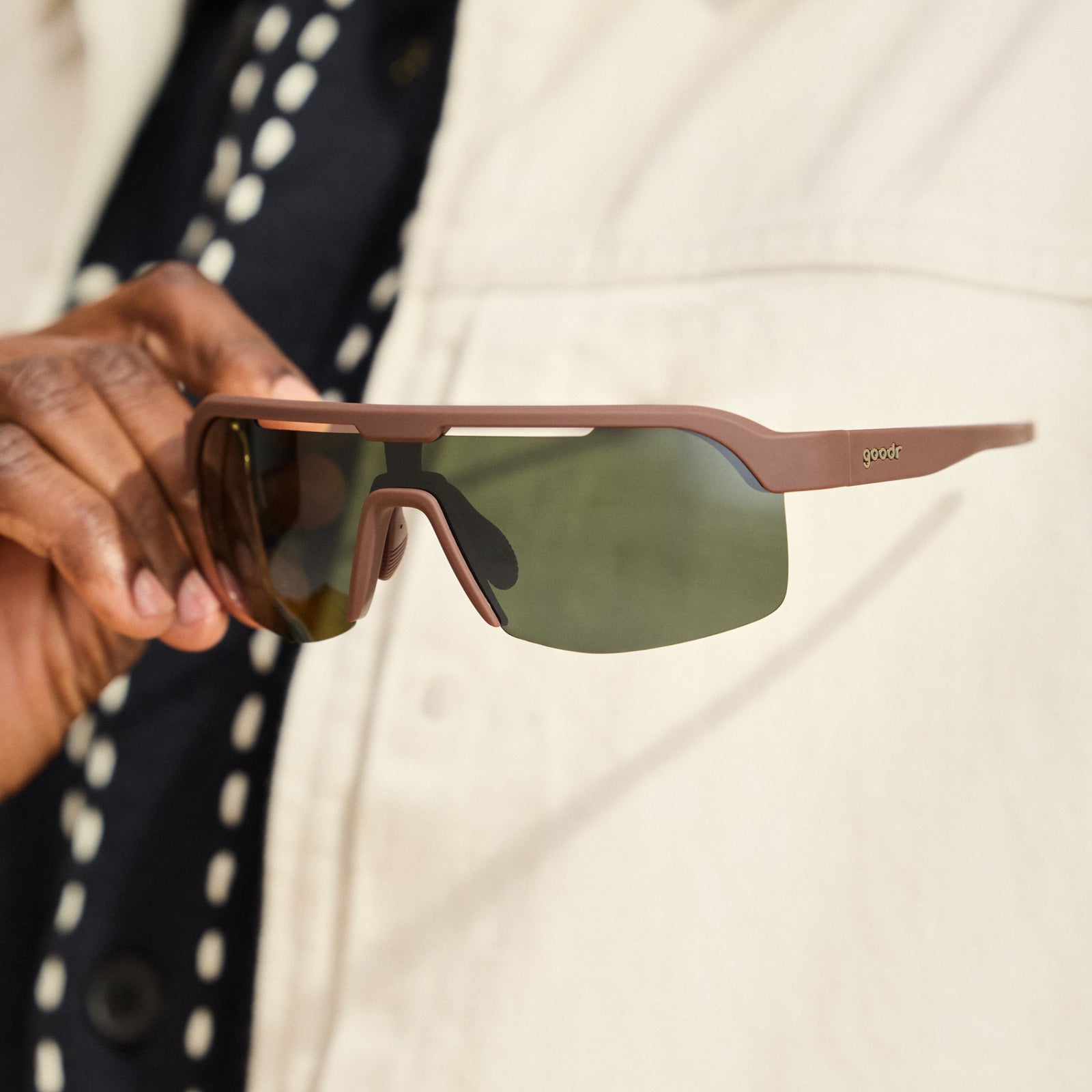 Close-up of a man’s hand holding a pair of brown Bolt G sunglasses in front of his chest. He wears a cream-colored jacket.