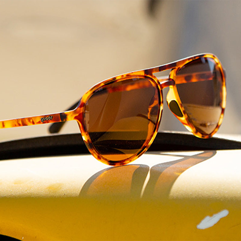 Three-quarter angle view of brown tortoiseshell aviator sunglasses with brown non-reflective lenses sitting on top of a yellow metal surface.