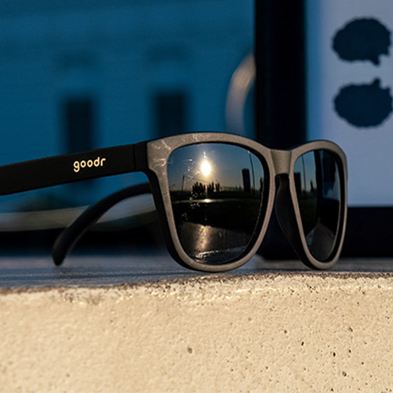 Side view of black sunglasses sitting on a concrete ledge outside with a city skyline reflected in their black lenses.