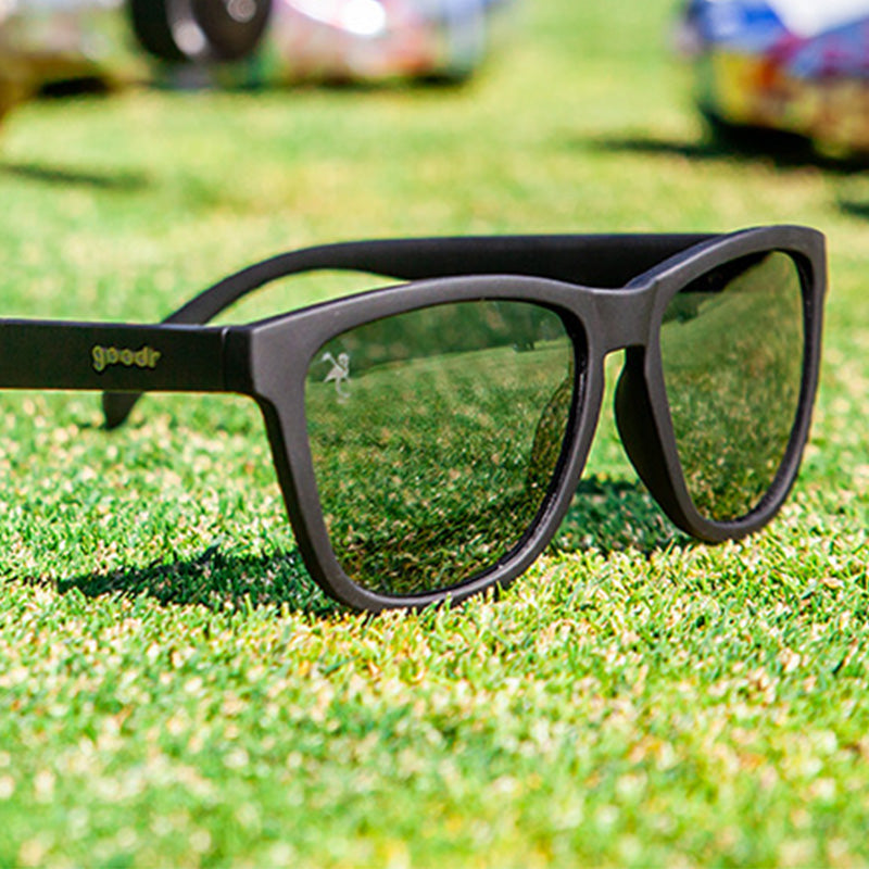 Three-quarter angle view of black sunglasses with black lenses sitting on the green turf of a golf course.