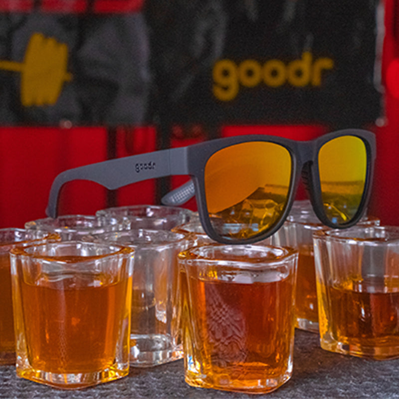 Three-quarter angle view of large square-shaped black sunglasses with mirrored amber lenses sitting on top of copious shot glasses filled with whiskey.