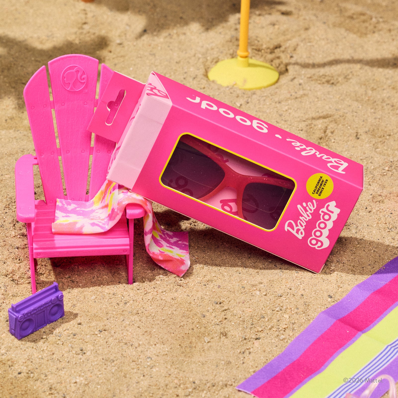 A custom pink Barbie™ sunglasses box is sitting in the sand surrounded by vibrant pink and purple Barbie beach accessories.