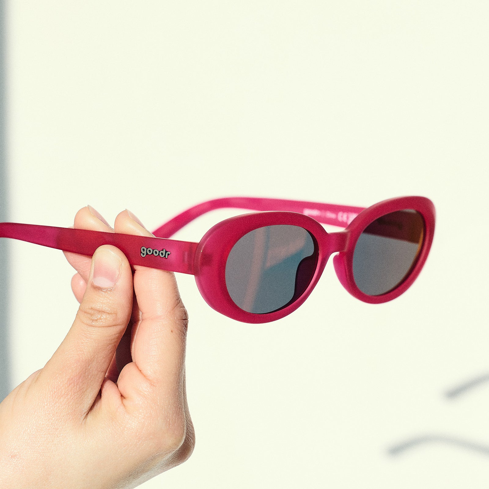 A manicured hand holds a pair of hot pink oval sunglasses with dark red polarized lenses in front of an off-white backdrop.