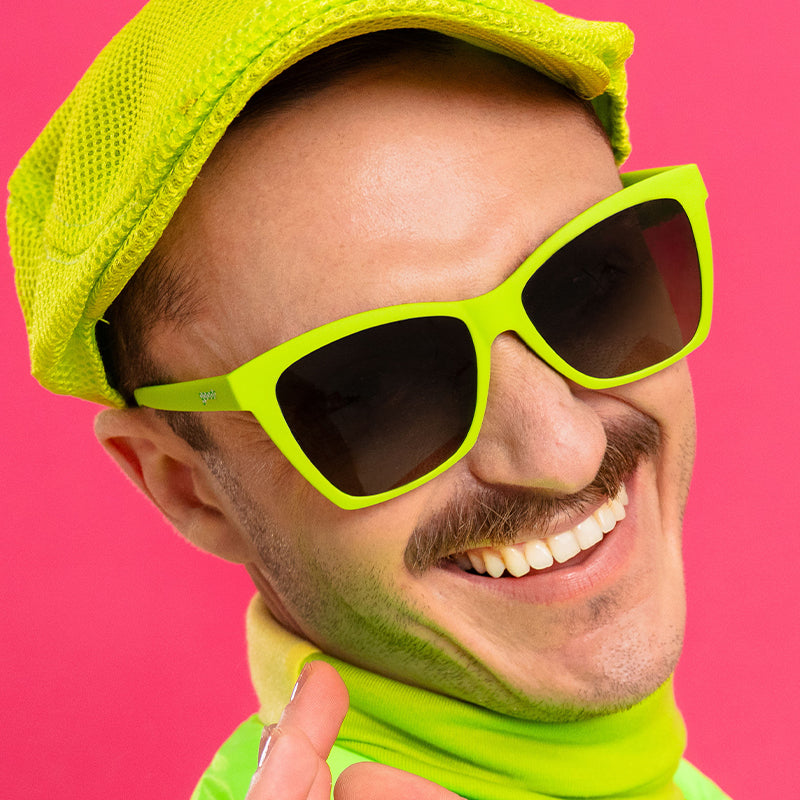 A man in a stylish neon green outfit smiles wearing bright green angled cat-eye sunglasses with black gradient lenses.
