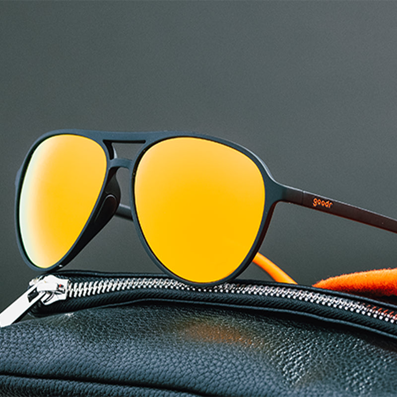 Three-quarter angle view of black aviator sunglasses with amber lenses sitting on top of a black leather bag.