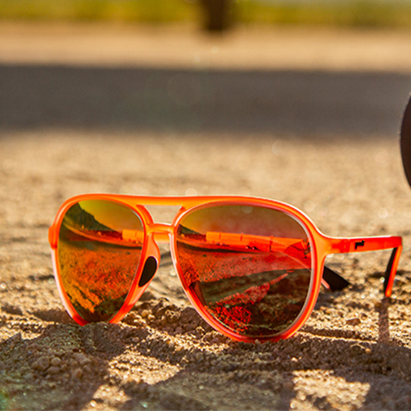 Three-quarter angle view of red aviator sunglasses with bright red reflective lenses sitting in sand along an airport tarmac.