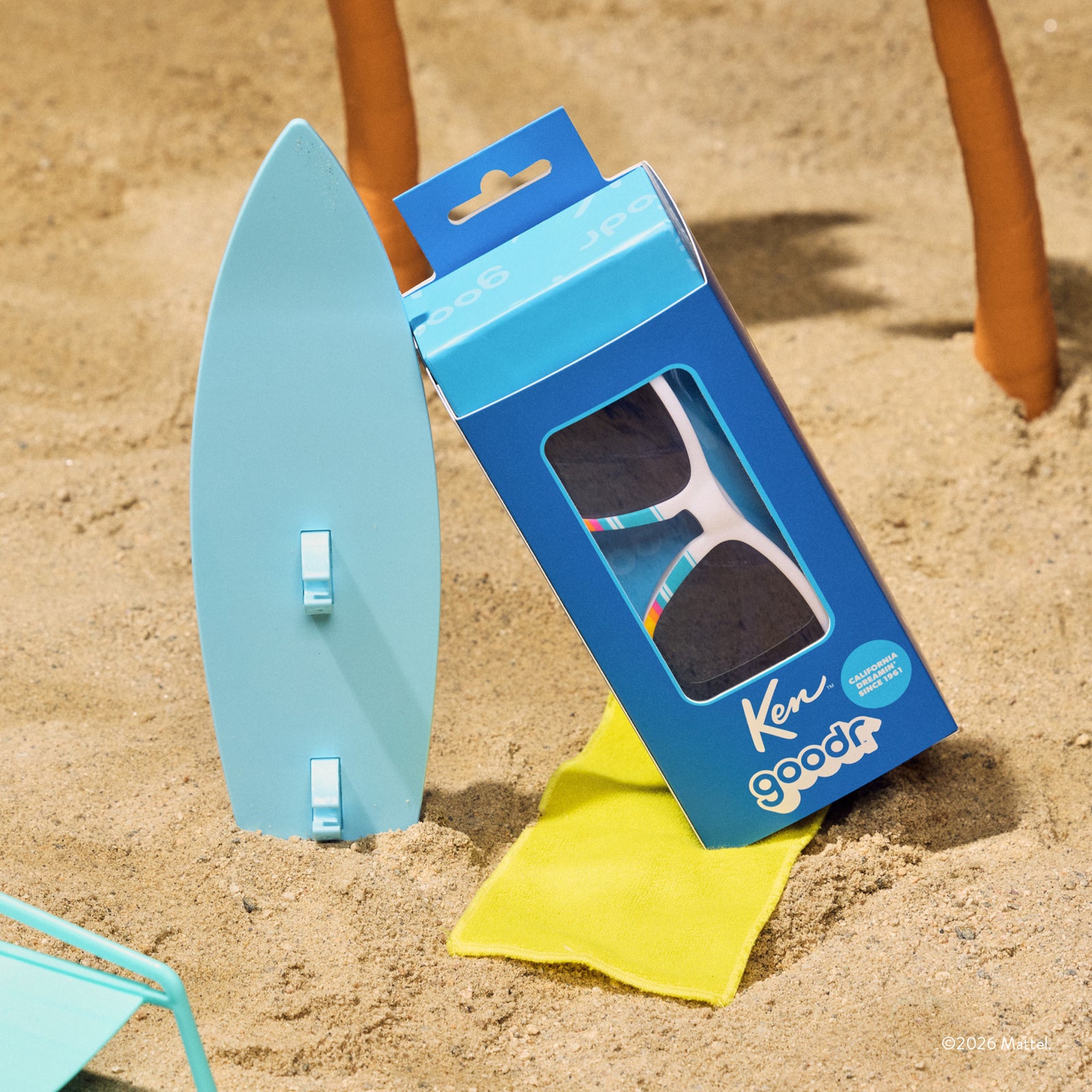 A custom blue Ken™ sunglasses box sits in the sand with a mini surfboard, showcasing the limited-edition beachy sunnies.