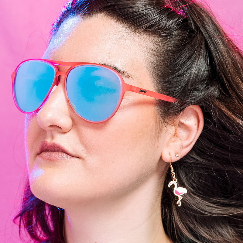 A woman in a pink room looks off the side wearing flamingo earrings and pink aviator sunglasses with mirrored reflective teal lenses.  