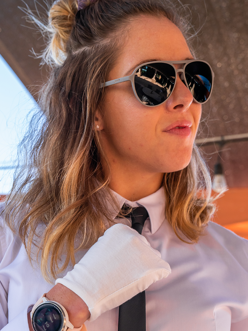 Woman wearing goodr sunglasses with black lenses, wearing a white button down, white gloves, and black tie