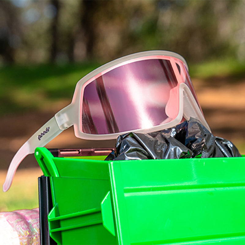 Three-quarter angle view of clear wraparound sunglasses with a rose-tinted single lens sitting atop a tiny toy dumpster.
