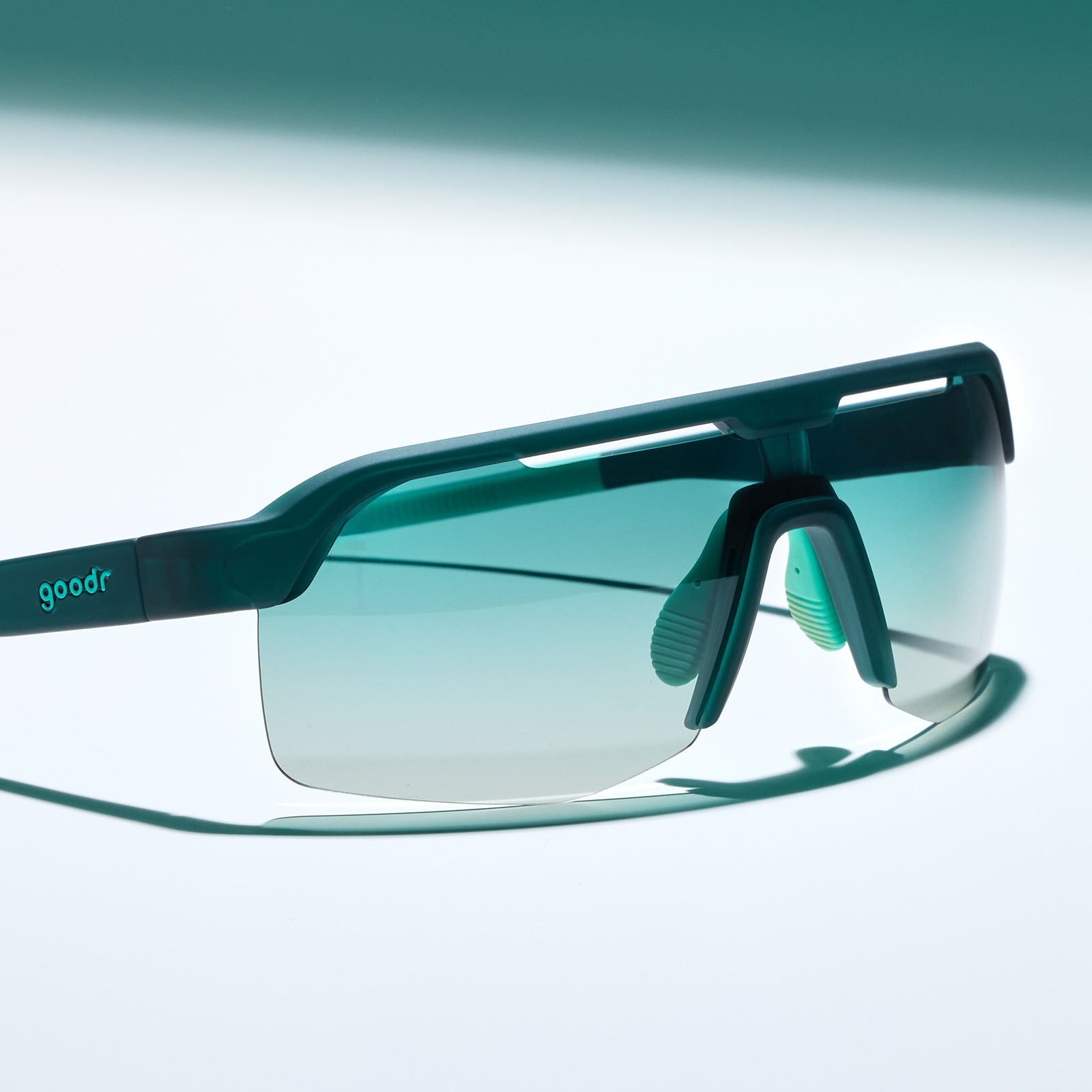 Three-quarter angle view of dark teal Bolt Gs with green gradient lenses sitting on a white surface.
