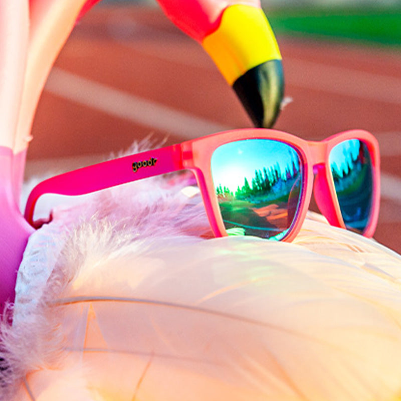 Three-quarter angle view of pink sunglasses with green reflective lenses sitting atop an inflatable flamingo-shaped pool floatie.