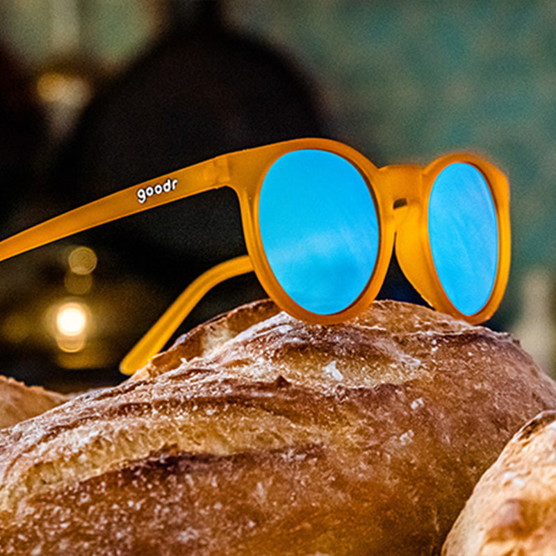 Three-quarter angle view of round orange sunglasses with light blue reflective lenses sitting atop a freshly baked loaf of bread.