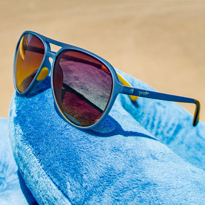 Three-quarter angle view of dark blue aviator sunglasses with dark amber lenses resting on a blue fuzzy travel neck pillow.