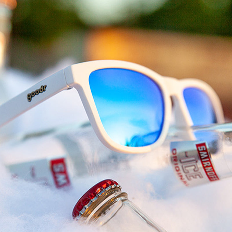 Three-quarter angle view of white sunglasses with blue reflective lenses sitting atop a bottled vodka soda in an ice bucket.