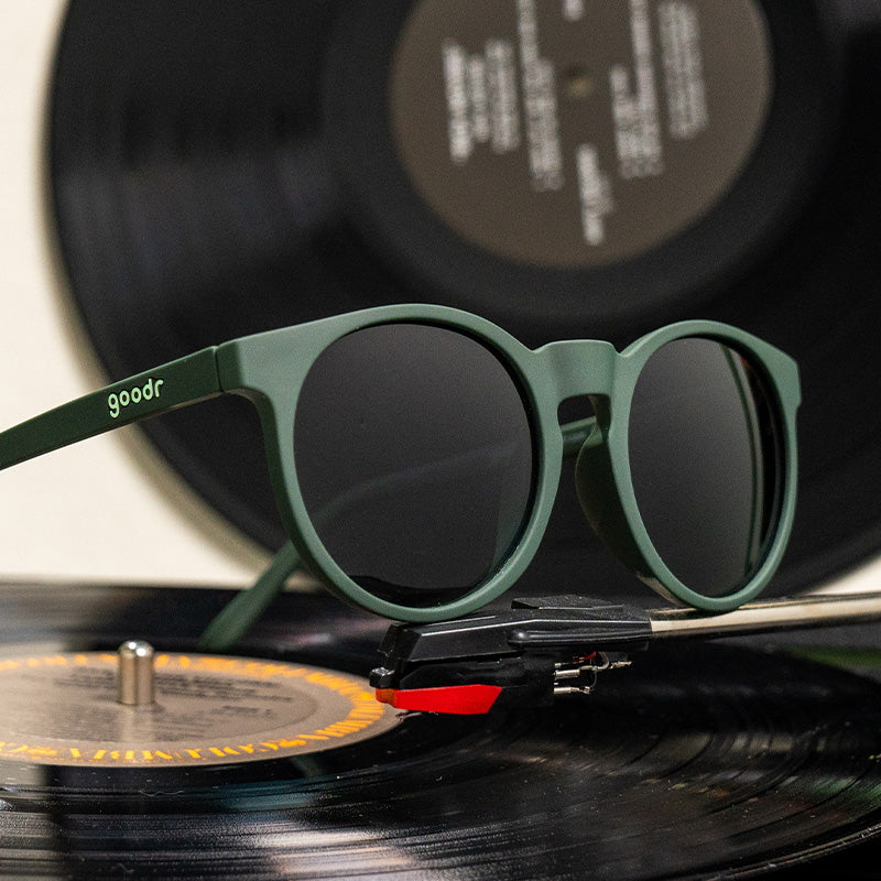 Three-quarter angle view of dark green round sunglasses with green circle-shaped lenses sitting atop a record player.