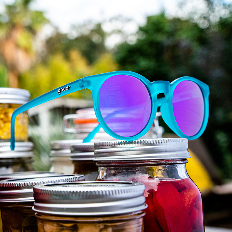Three-quarter angle view of round teal sunglasses with reflective purple lenses sitting atop mason jars of pickled veggies.