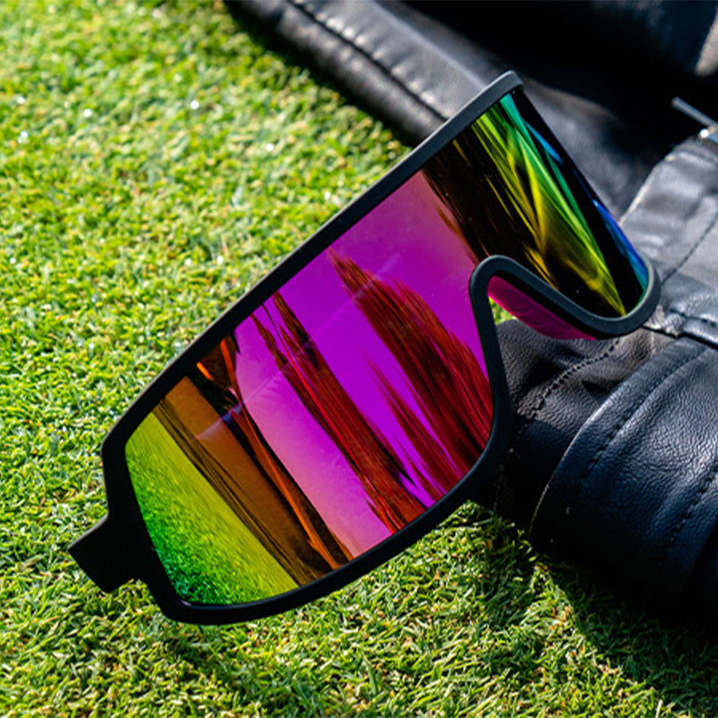 Three-quarter angle view of black wraparound sunglasses with a hot pink reflective lens sitting on a leather jacket on turf.