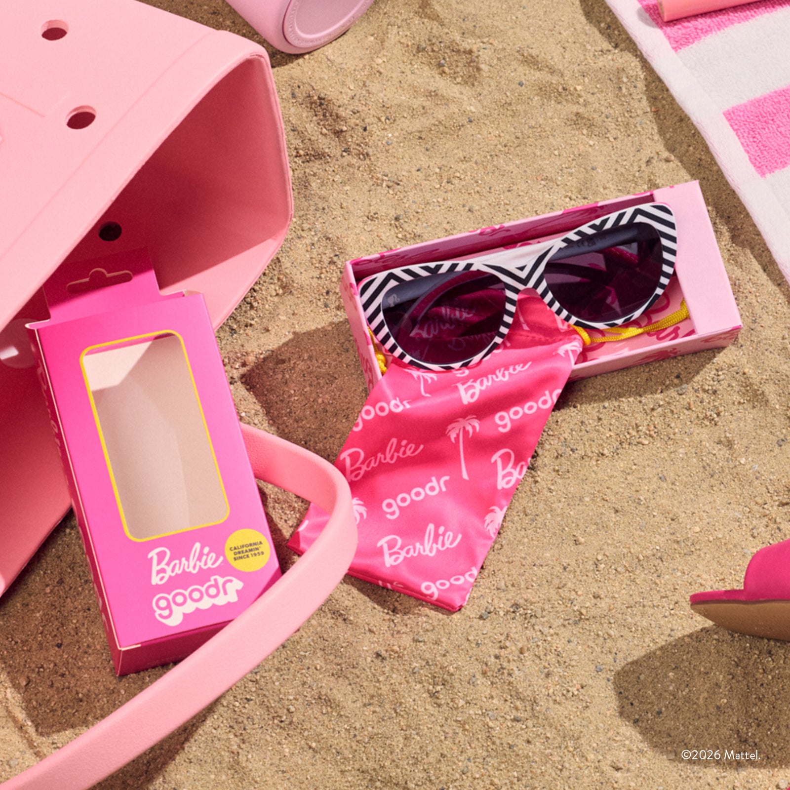 A custom pink Barbie™ sunglasses box sits open in the sand, showcasing the black-and-white sunnies and the hot pink pouch.