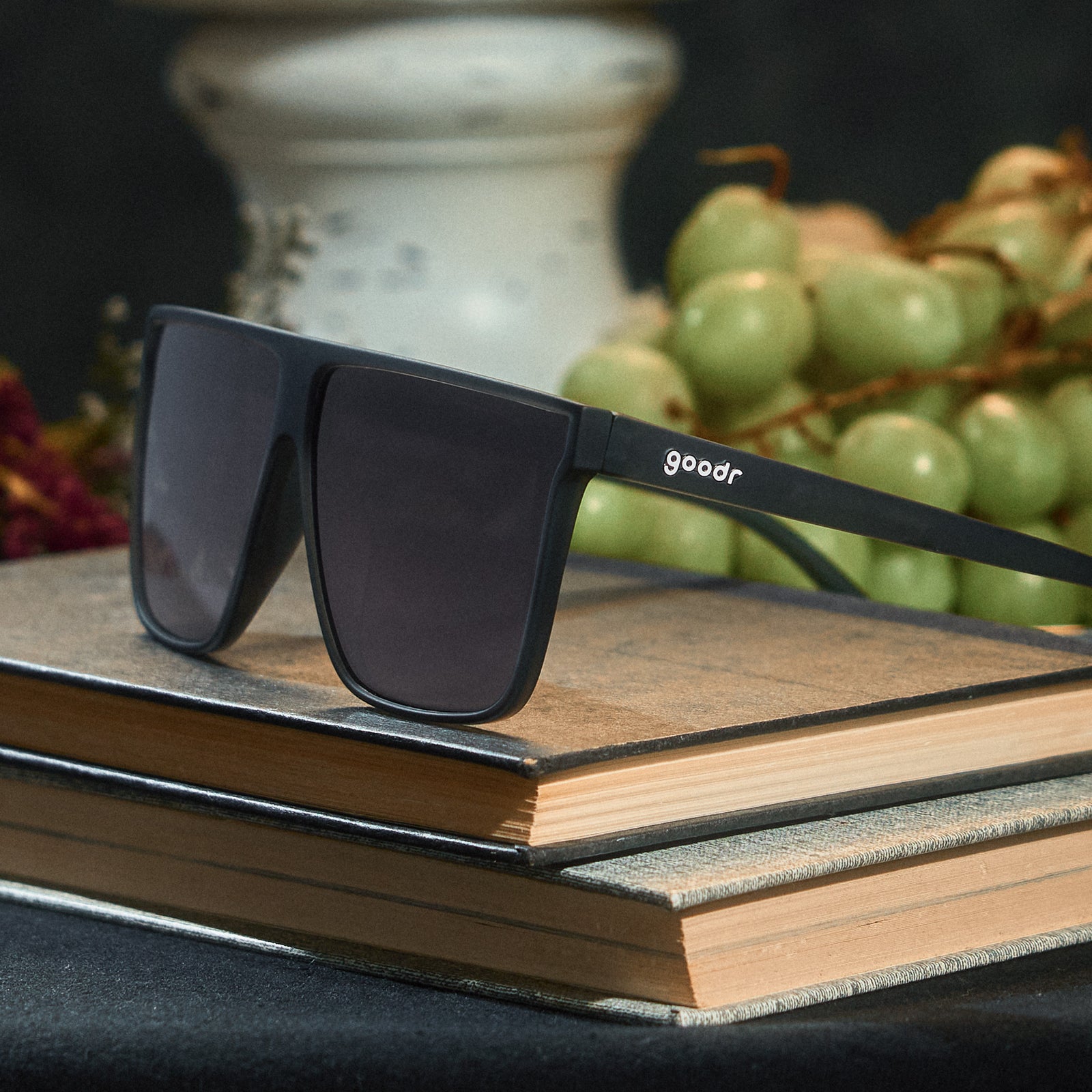 A pair of translucent gray, flat-top Fly G sunnies sits atop some vintage books, a bunch of green grapes in the background.