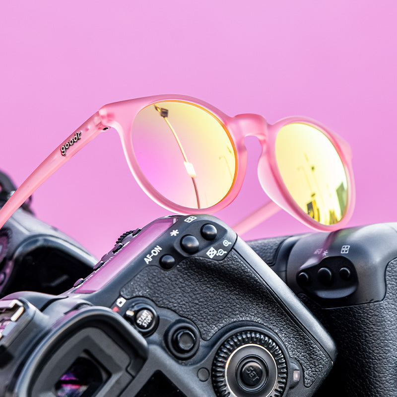 Three-quarter angle view of round pink sunglasses with pink reflective lenses sitting atop a pile of cameras.
