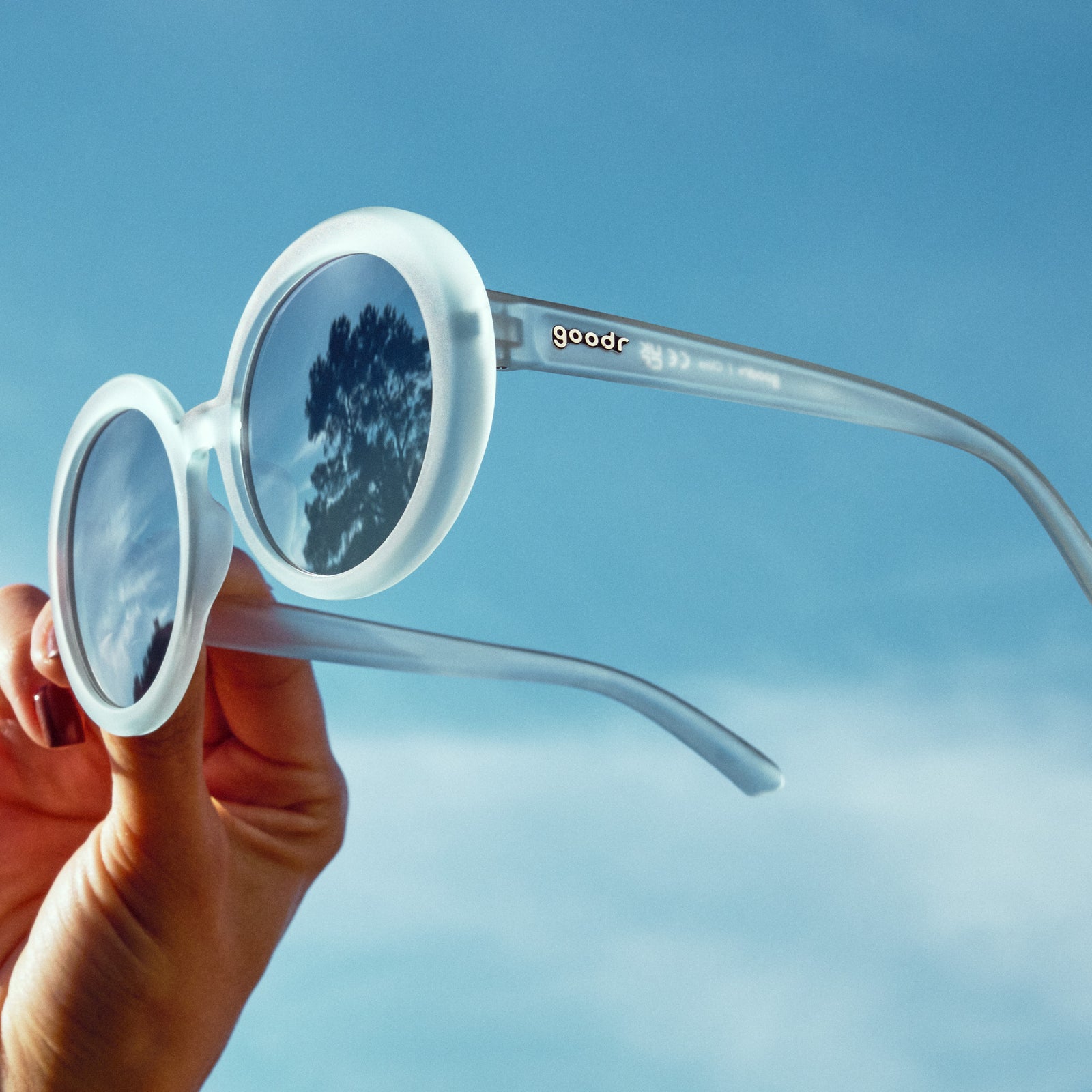 A hand holds up a pair of oversized round sunglasses with translucent blue frames and silver reflective gradient lenses against the sky.