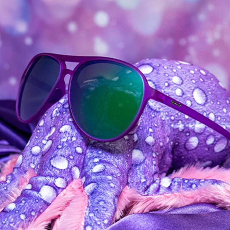 Three-quarter angle view of purple aviator sunglasses with green reflective lenses ar positioned on a purple stuffed octopus.