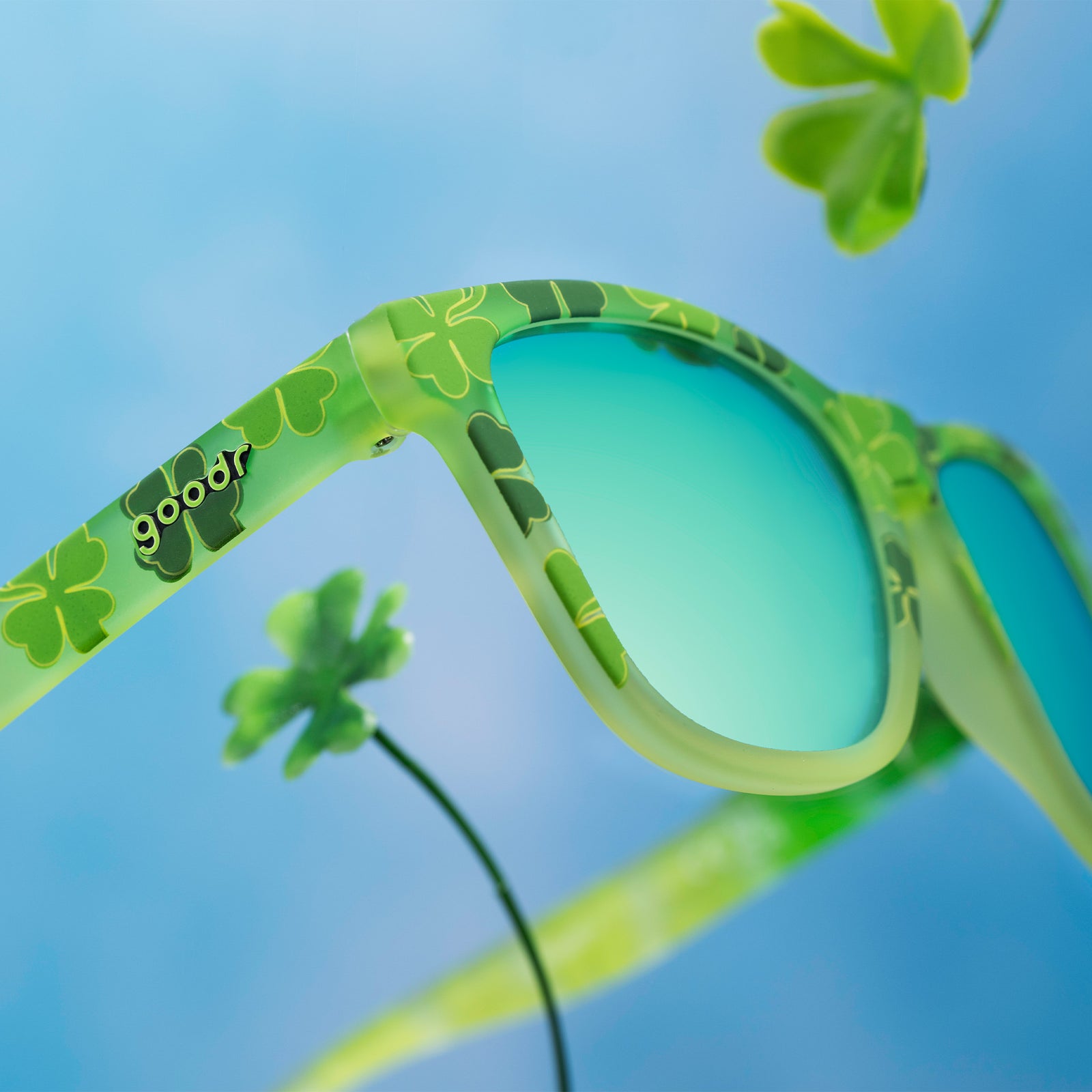 Three-quarter angle close-up view of green shamrock print shades w/ green+gold reflective lenses floating in the sky.