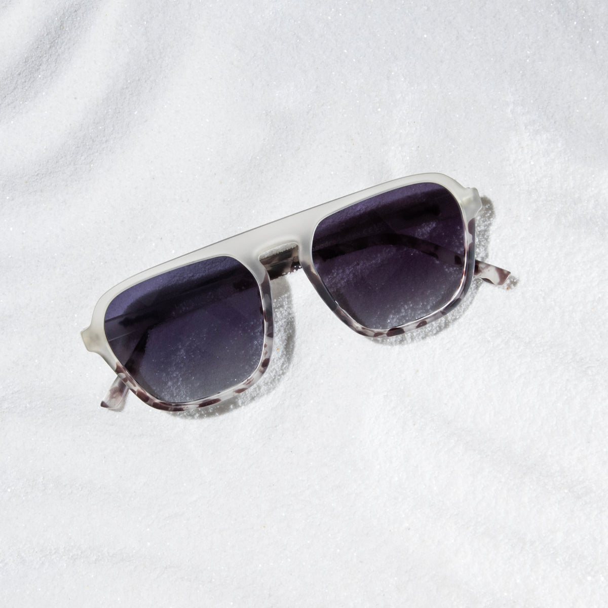 Translucent light gray retro aviator sunglasses with the goodr logo resting on a bed of snow.