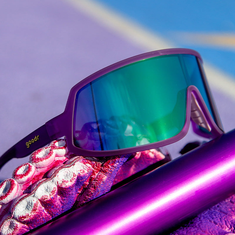 Three-quarter angle view of wraparound sunglasses with a purple frame and green lens sitting on a rubber tentacle glove.