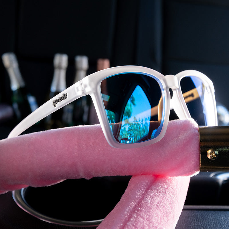 Three-quarter angle view of clear sunglasses with reflective blue lenses sit atop pink velvet ropes, champagne behind it.