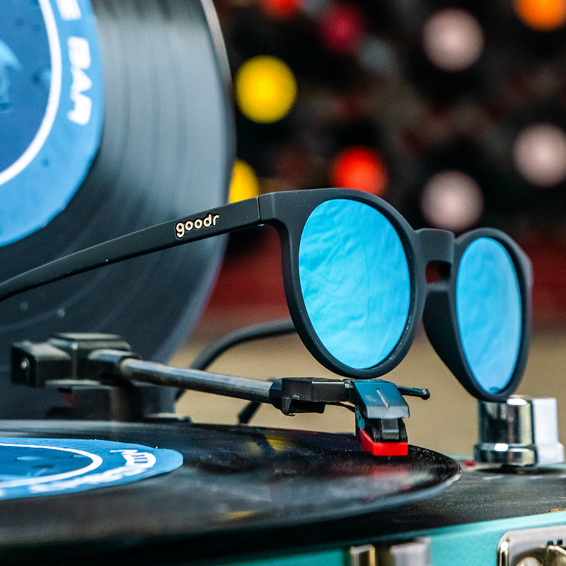 Three-quarter angle view of black round sunglasses with reflective blue circle-shaped lenses sitting atop a record player.