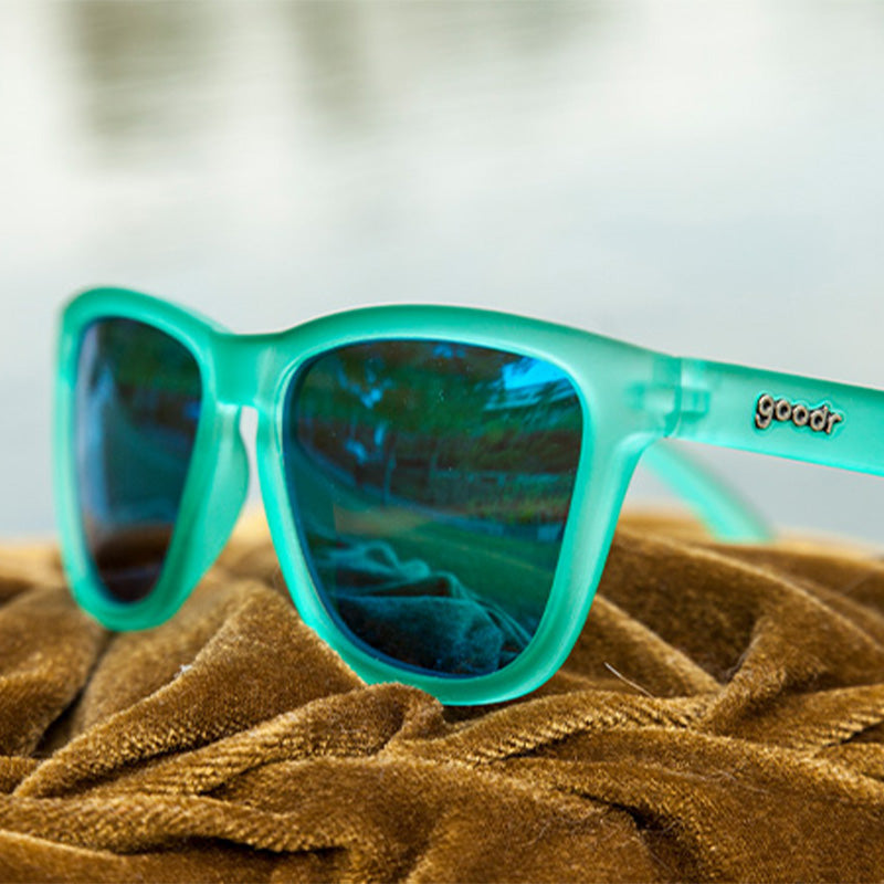 Three-quarter angle view of square teal sunglasses with teal reflective lenses sitting atop a pile of gold velvet fabric.