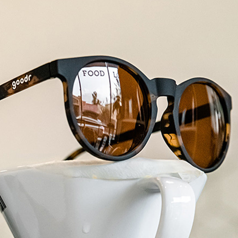 Three-quarter angle view of round brown sunglasses with brown non-reflective lenses sitting atop a pour-over coffee dripper.