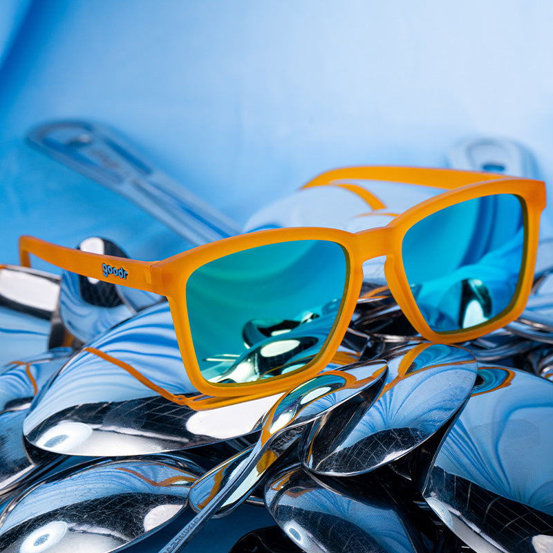 Three-quarter angle view of small square-shaped orange sunglasses with mirrored reflective lenses placed atop a bed of spoons.