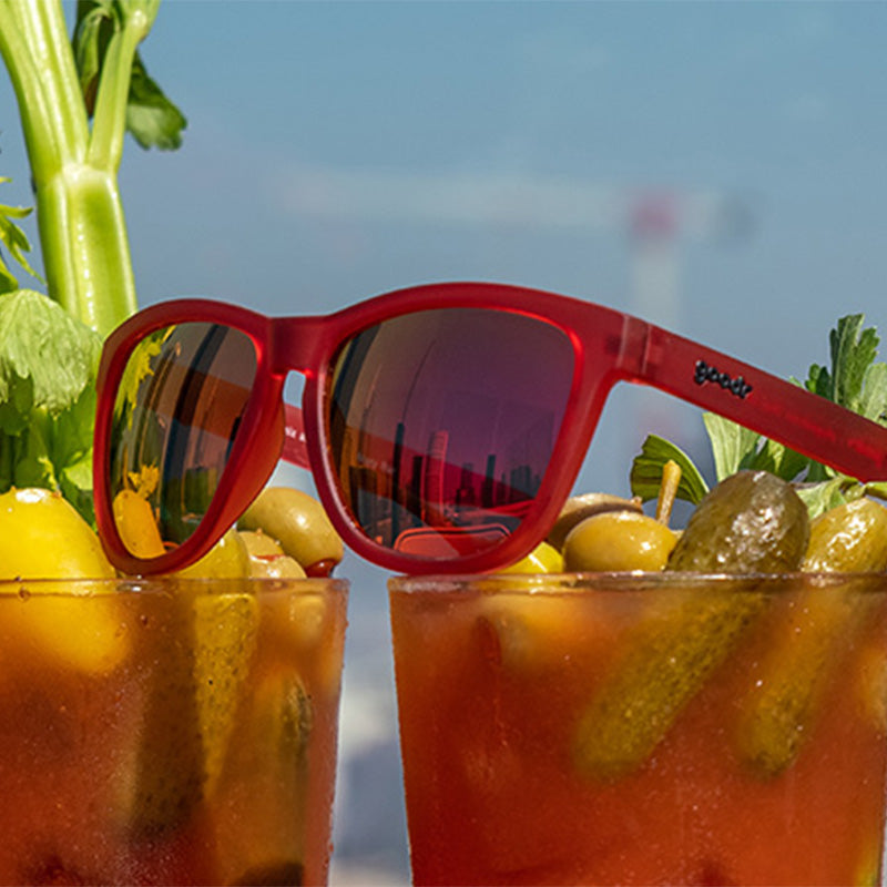 Three-quarter angle view of red sunglasses with red reflective lenses sitting atop two bloody mary cocktails.