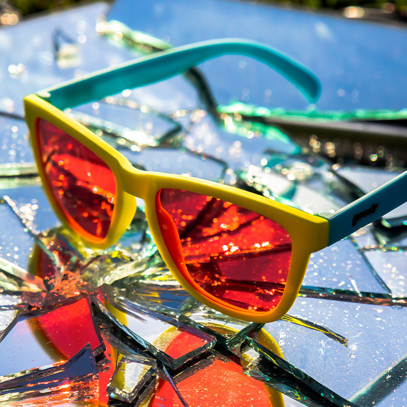 Three-quarter angle view of yellow and pale blue sunglasses with rose lenses sitting atop a shattered mirror.